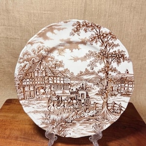 May include: A decorative plate with a brown transferware design depicting a horse-drawn carriage, buildings, and trees. The plate is white with a brown border and is displayed on a wooden surface.