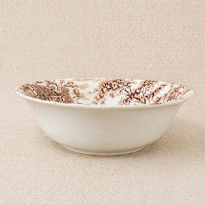 May include: A white ceramic bowl with a scalloped rim. The interior rim features a brown transferware design of trees and a building. The bowl has a simple, classic design.