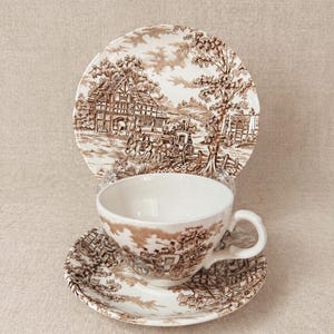 Könnte beinhalten: Ein Vintage-Teeservice mit braunem und weißem Transferdruck. Das Set umfasst eine Teetasse, eine Untertasse und einen Teller, alle mit einer detaillierten ländlichen Szene. Die Tasse und Untertasse sind vor dem Teller platziert.
