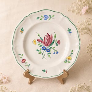 May include: A round ceramic plate with a scalloped edge and a green painted rim. The center features a hand-painted floral motif with a large central red flower surrounded by smaller blue and yellow blossoms and green leaves on a white background.
