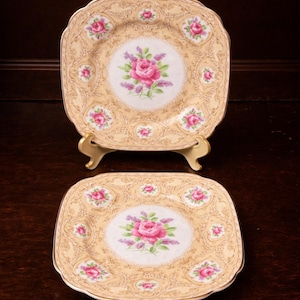 Op de afbeelding: Twee vierkante borden met een bloemmotief. De borden hebben een crèmekleurige achtergrond met een roze roos en een lavendelcentrum. De borden zijn afgewerkt met goud en worden gepresenteerd op een donkere houten ondergrond.