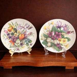 Könnte beinhalten: Zwei dekorative Teller mit Blumendesigns. Jeder Teller zeigt einen Strauß bunter Blumen, darunter Iris, Tulpen und andere Blüten, vor einem cremefarbenen Hintergrund. Die Teller werden auf dekorativen Ständern präsentiert.