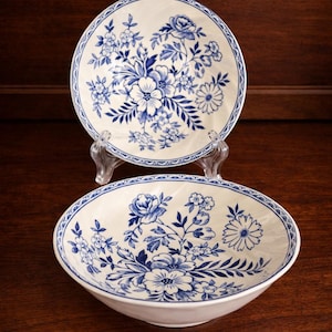 Może przedstawiać: Dwie ceramiczne miski z kremową podstawą i niebieskimi wzorami kwiatowymi. Miski zdobione są szczegółowym niebieskim wzorem kwiatowym i niebieską obwódką. Jedna miska jest okrągła, druga owalna.