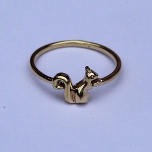 以下が含まれることがあります： 金色のリングで、リスのデザインが特徴です。リングは細いバンドと、カールした尾を持つ詳細なリスの姿をしています。リスは右を向いています。リングは白い背景にあります。