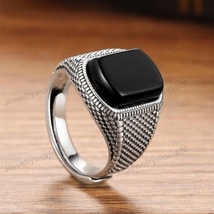 Puede incluir: Anillo plateado con una piedra cuadrada de ónix negro. El anillo tiene una banda texturizada con un patrón detallado. Se muestra sobre una superficie de madera.