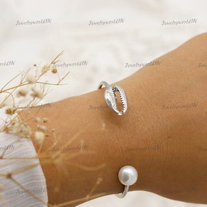 Puede incluir: Pulsera de plata con un dije de concha marina y un acento de perla. La pulsera está en la muñeca de una persona.