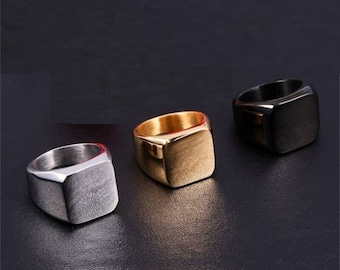May include: Three square signet rings in different colors: silver, gold, and black. The rings are all made of metal and have a smooth, polished finish.