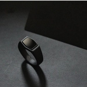 Pode incluir: Um anel preto com uma pedra preta retangular embutida em uma faixa preta fosca. O anel tem um design geométrico moderno e é exibido em uma superfície escura. A sombra do anel é visível.