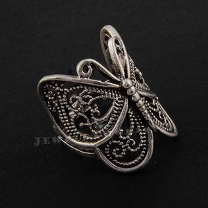 Peut inclure: Broche argentée en forme de libellule, avec des détails de filigrane complexes sur les ailes et le corps. La broche a un design en trois dimensions, avec les ailes légèrement relevées. La libellule est posée sur un fond sombre.
