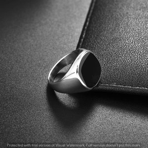 Peut inclure: Une bague en argent poli avec une grande pierre d'onyx noire carrée. La surface polie de la bague reflète la lumière, sur un fond noir texturé. Le design est simple et élégant, adapté au quotidien.