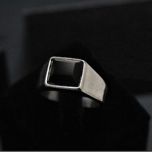 Pode incluir: Um anel de prata com uma pedra preciosa preta quadrada engastada em um engaste de bezel.