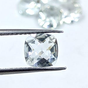 May include: A clear, faceted, cushion-cut gemstone held by tweezers. The gemstone is a light, pale, transparent colour.