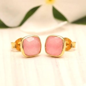 May include: A pair of stud earrings featuring square-cut, faceted pink stones set in polished gold-tone metal. Each earring has a smooth, rounded square bezel frame that secures the light pink gemstone, complete with standard gold-tone earring backs.