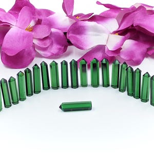 Op de afbeelding: Een rij van 20 groene edelsteenpunten die in een halve cirkel op een wit oppervlak zijn gerangschikt. De punten zijn allemaal even groot en even gevormd, en ze zijn allemaal puntig aan de bovenkant. Er is één punt in het midden van de halve cirkel dat iets kleiner is dan de andere.