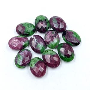 Puede incluir: Un conjunto de gemas ovaladas de rubí y zoisita. Las piedras muestran una mezcla de colores rojo intenso y verde, con superficies facetadas que capturan la luz. Se presentan sobre un fondo blanco.