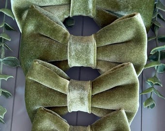 May include: Four sage green velvet bows arranged in a vertical stack, each featuring a central fabric knot and pleated wings. The bows are crafted from a soft, light-reflecting velvet material in a consistent olive-toned green shade throughout.