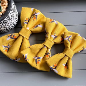 May include: Three yellow bow ties with a bee pattern. The fabric bow ties feature a repeating pattern of bees in black, orange, and white. The bow ties are arranged on a grey surface, with a decorative plant pot in the background.
