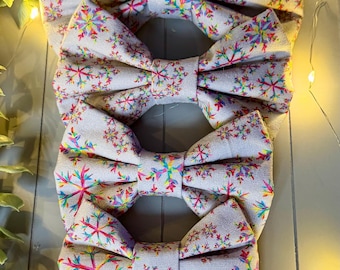 May include: A stack of four bow ties made from a light gray fabric featuring a repeating pattern of colorful, multi-hued snowflake designs in shades of pink, blue, yellow, and green. Each bow tie has a central fabric knot detail.