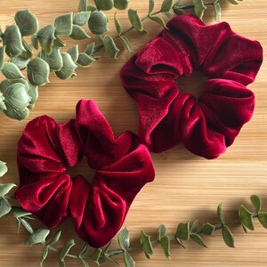 May include: Two burgundy velvet scrunchies are displayed on a wooden surface, with green eucalyptus leaves. The scrunchies are made of a soft, plush material and are a deep red color.