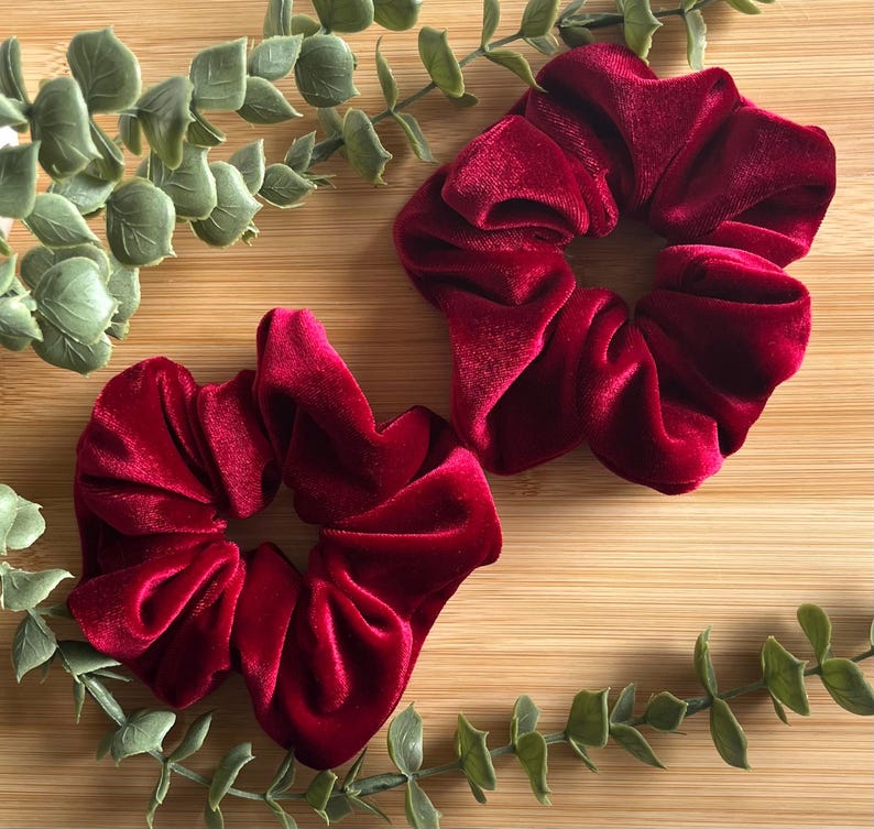 May include: Two burgundy velvet scrunchies are displayed on a wooden surface, with green eucalyptus leaves. The scrunchies are made of a soft, plush material and are a deep red color.