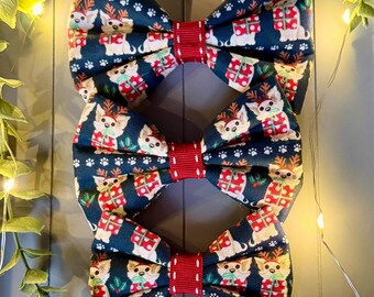 May include: Three fabric bow ties featuring a dark blue background with a repeating pattern of small dogs wearing reindeer antlers and holding red and white gift boxes, accented by a central red ribbed fabric band with white stitching.