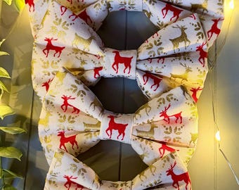 May include: A set of four fabric bow ties in varying sizes, featuring a repeating pattern of red and gold reindeer silhouettes and gold snowflake motifs on a cream-colored background, arranged vertically in a stack.