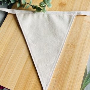 May include: A plain, cream-coloured fabric pennant flag. The triangular flag has a stitched border and a fabric strip for hanging. It is displayed on a light brown wooden surface, with greenery in the background.