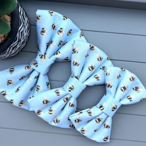 May include: Three light blue bow ties featuring a repeating pattern of black and yellow bees. The bow ties are arranged in a cluster, with one larger and two smaller sizes. The background is a grey surface.
