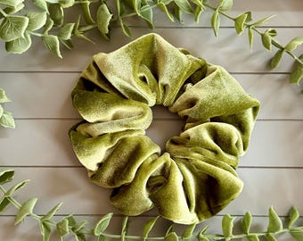 May include: A circular hair scrunchie made of olive green velvet fabric. The accessory features a gathered, pleated texture with a soft, light-reflecting sheen across the surface of the material, creating a voluminous and rounded shape.