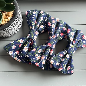 May include: Three navy blue fabric bow ties with a floral pattern of white and pink flowers. The bow ties are arranged on a grey surface, with a potted plant in the background.