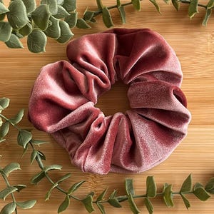 May include: A dusky pink velvet scrunchie, a hair accessory, is the focal point of the image. The scrunchie is round and gathered, with soft folds. Green leafy sprigs are arranged around the scrunchie on a wooden surface.