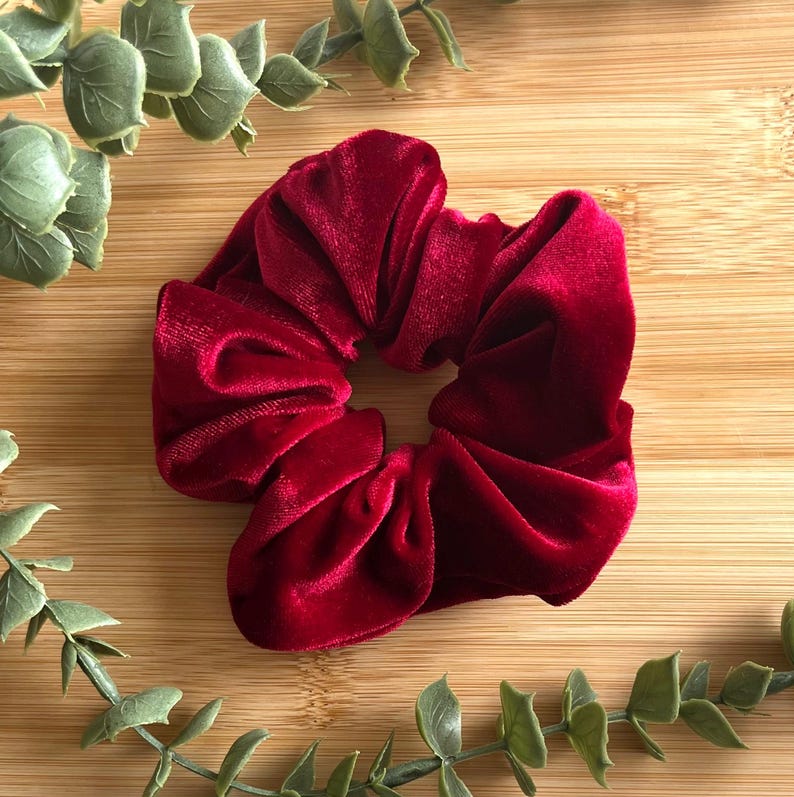 May include: A deep red velvet scrunchie, a hair accessory, sits on a wooden surface. The scrunchie is made of a soft, plush material and is gathered in a circular shape. Green leafy sprigs frame the scrunchie.