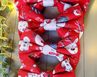 May include: A stack of four red fabric bow ties featuring a repeating Christmas-themed print. The pattern includes illustrations of reindeer, gingerbread men, candy canes, and ornaments in shades of white, gray, and red across the fabric.