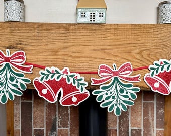May include: A festive garland featuring alternating die-cut shapes of red glittery bells and green mistletoe sprigs with red bows, all strung together on a thin red ribbon. The shapes are outlined in white and display a vibrant holiday color palette.