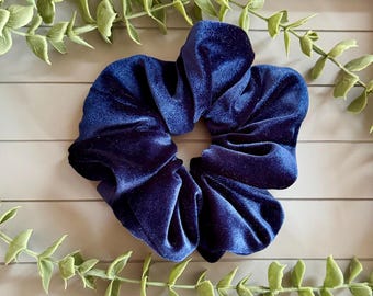 May include: A deep navy blue velvet scrunchie with a soft, plush texture and gathered fabric design. The hair accessory features a circular shape with a central opening, showcasing the rich, dark blue color and the light-reflecting velvet material.