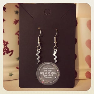 May include: Silver lightning bolt earrings displayed on a black card. The earrings are suspended from silver hooks. The card features a circular sticker with the text: "Handmade by Kay. Find us on Etsy... Sparkle & Glimmer."