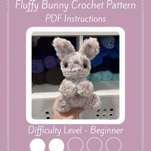 Op de afbeelding: Een zacht, grijs gehaakt konijn staat centraal in deze afbeelding. Het konijn heeft een gedetailleerd gezicht en oren. De tekst "Fluffy Bunny Crochet Pattern PDF Instructions" staat bovenaan. De moeilijkheidsgraad is beginner.