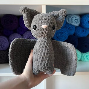 May include: A handmade gray crochet bat with large black eyes and a small pink nose. The bat has large, textured wings and ears. The plush toy is held in a hand, with shelves of colorful yarn in the background.