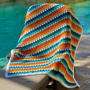 May include: A vibrant, hand-crocheted blanket draped over a chair. The blanket displays a striped pattern with orange, teal, white, and yellow colours. The edges have a white border. The blanket is approximately 152 cm by 127 cm.