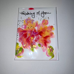 May include: A white card with the words "Thinking of you" written in black ink. The card features a watercolor painting of pink, orange, and yellow flowers with gold glitter accents.