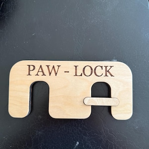 May include: A wooden "PAW-LOCK" device, designed with a rectangular shape and cut-out sections. The text "PAW-LOCK" is laser-etched across the top. The device is made of light-colored wood and sits on a dark surface.