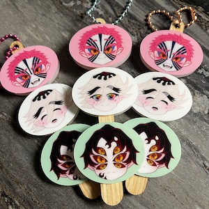 May include: Eight round, colorful, printed, paper,  popsicle stick style,  keychains with anime characters.  The characters have black hair and different colored eyes.  The keychains are on a wooden surface.