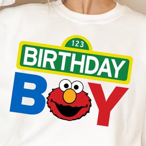 May include: White crew neck sweatshirt with a colorful graphic. The design features the words "BIRTHDAY BOY" in blue, red, and green, with a cartoon character in the center. The sweatshirt has long sleeves.