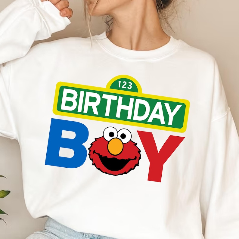 May include: White crew neck sweatshirt with a colorful graphic. The design features the words "BIRTHDAY BOY" in blue, red, and green, with a cartoon character in the center. The sweatshirt has long sleeves.