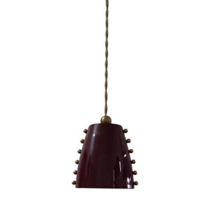 Op de afbeelding: Bordeauxkleurige hanglamp met een conische kap. De kap is versierd met kleine, gelijkmatig verdeelde messingkleurige klinknagels. Het licht hangt aan een gedraaid, goudkleurig snoer en heeft een messingkleurige bovenkant.
