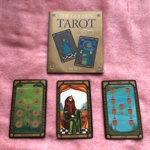 May include: Three tarot cards, the Nine of Pentacles, the King of Wands, and the Nine of Cups, laid out on a pink surface. The cards are from the Golden Tarot deck by Liz Dean.