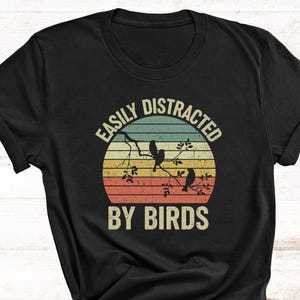 May include: Black t-shirt with a retro design featuring a sunset, birds on a branch, and the text "EASILY DISTRACTED BY BIRDS". The design is in shades of orange, yellow, green, and blue.