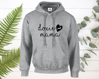 Op de afbeelding: Een lichtgrijze hoodie met een voorzak en een trekkoord. De hoodie toont de woorden "doxie mama" in een scriptlettertype, met een zwart hart en een silhouet van een teckel.