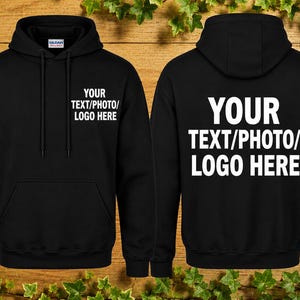 Puede incluir: Sudadera con capucha negra con bolsillo delantero y cordón. La parte delantera y trasera de la sudadera con capucha presentan el texto "YOUR TEXT/PHOTO/LOGO HERE" en blanco. La sudadera con capucha se muestra sobre un fondo de madera con hojas verdes.