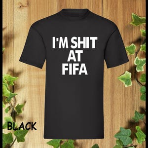 Puede incluir: Camiseta negra con texto blanco que dice "I'M SHIT AT FIFA".